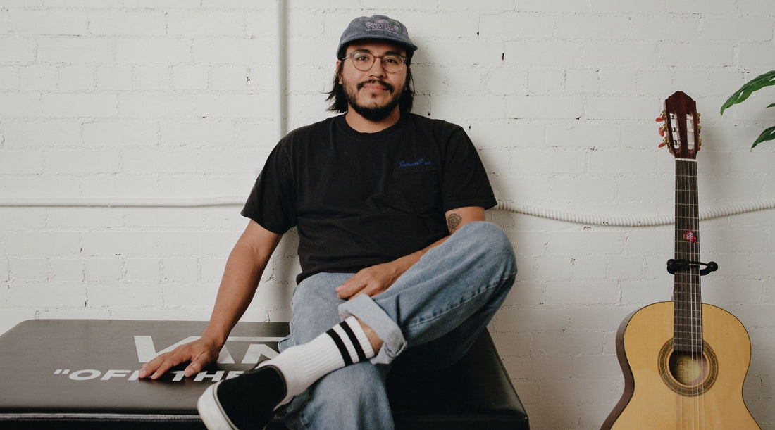 A person, Tony Perez, sits on a black surface next to a guitar with his leg crossed over his knee. One arm rests on his lap, while the other rests at his side. He wears a black shirt with jeans, and a grey baseball cap.