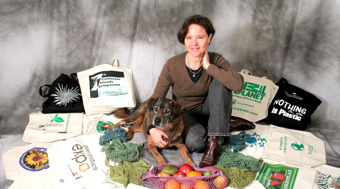 Sharon Rowe, a woman with short brown hair and a light complexion, sits among an array of printed ECOBAGS totes and string bags with her brown dog, Hudson. 