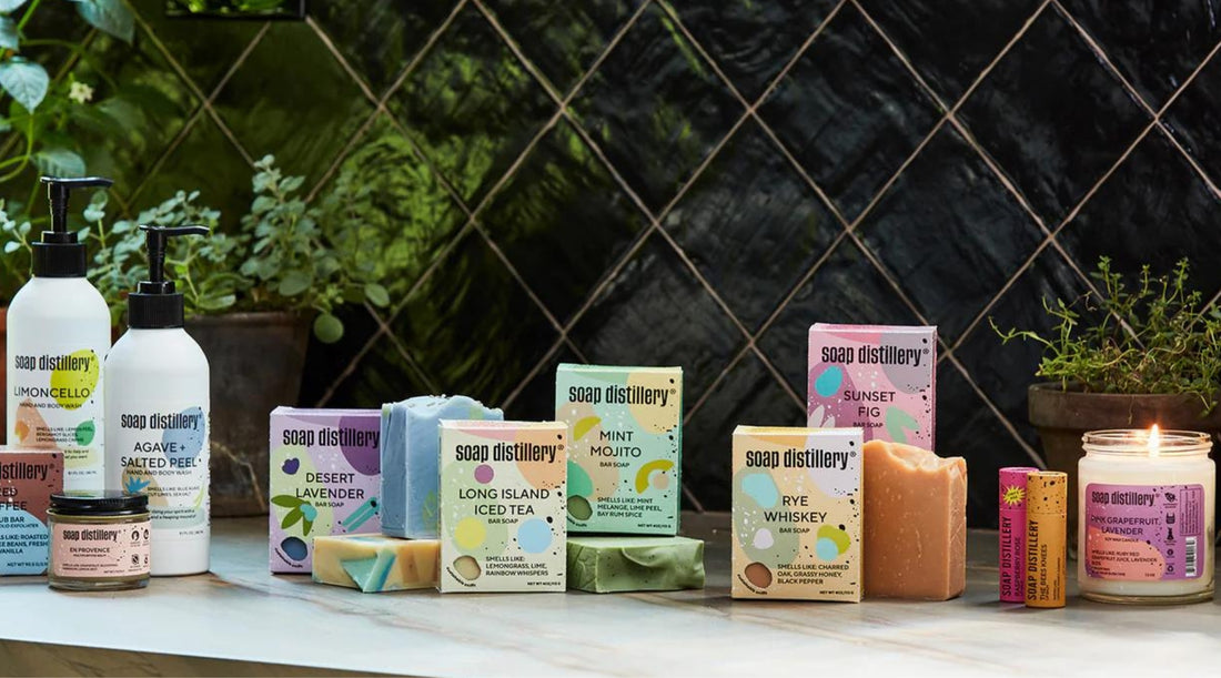 An array of Soap Distllery soaps, balms, and candles are against a black tiled backsplash, sitting on a white marble countertop.